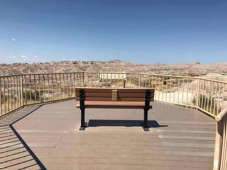 A bench on a boardwalk faces a badlands landscape extending into the horizon under a blue sky.