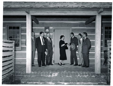 Woman in a black dress stands in front of a cabin door, three men in suits stand on each side of her