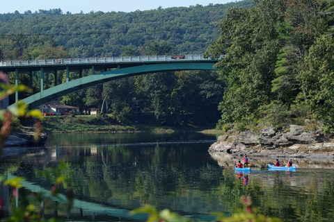 A set of 2 canoes with 2 paddlers in each canoe paddle towards the Narrowsburg Bridge.