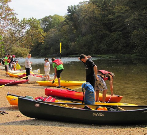a group of young people on shore next to river with canoes and kayaks