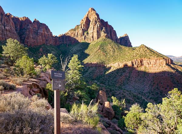 View from a sandy overlook of a triangular sandstone mountain with green foliage on the foothills.