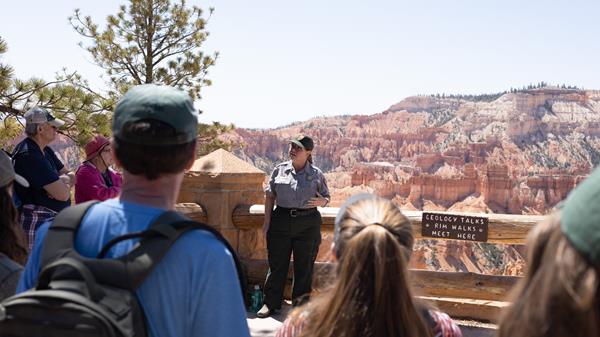 A ranger in uniform stands in front of a crowd with a background of red rock formations