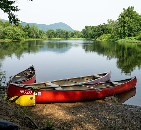 Three canoes sit at the dirt take-out of a campsite. The landscape reflects on the river.