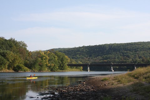 person in yellow kayak paddles in river with aqueduct-converted bridge in background.