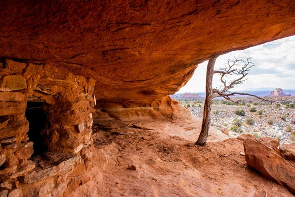 A stone and clay structure sits underneath an orange sandstone alcove on a cloudy day.