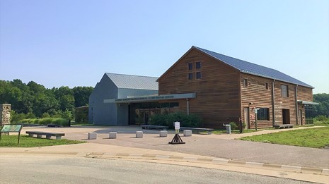 A building with three pitched roofs and a flat roof, surrounded by grass and trees in the background