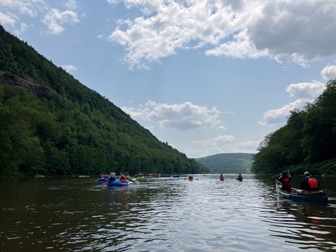 Group of rafts and canoes paddle on river in between forested two mountains/hills.