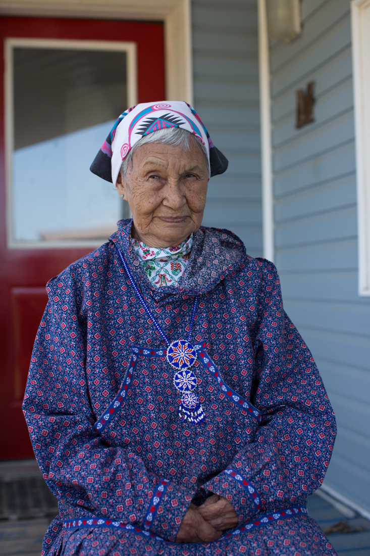 A woman sits on porch in full regalia with headscarf, kuspuk and rosette beaded necklace.