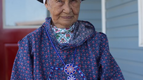 A woman sits on porch in full regalia with headscarf, kuspuk and rosette beaded necklace.