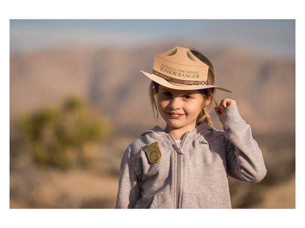 A young girl wearing a Junior Ranger badge and hat with mountains and a Joshua tree in the distance.