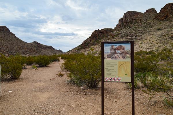 A metal sign with stands to the side of a gravel trail, with hills rising up around the trail.