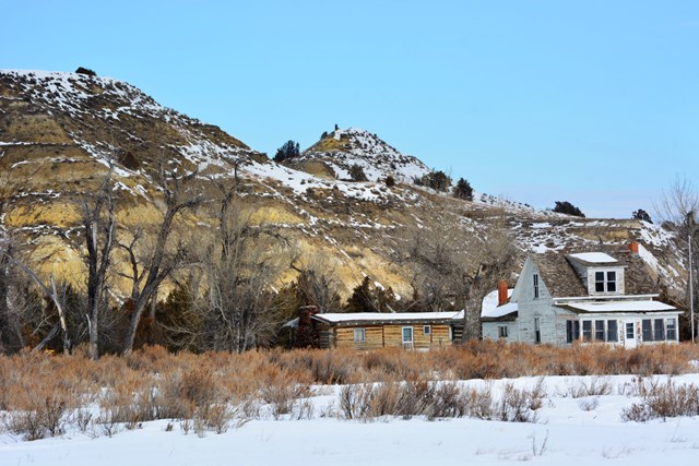 A white ranch house beside a log building in winter, with trees and buttes behind