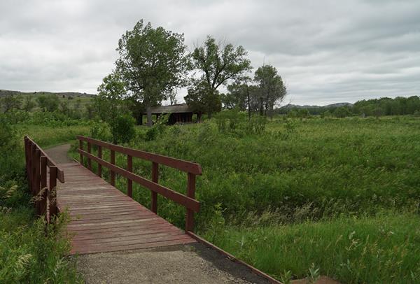 A paved trail leads to a bridge over a creek. A picnic shelter is seen behind some trees.