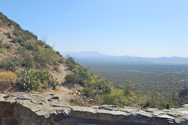 Rock wall along the bottom of the frame with a trail climbing a mountain to the left above a valley