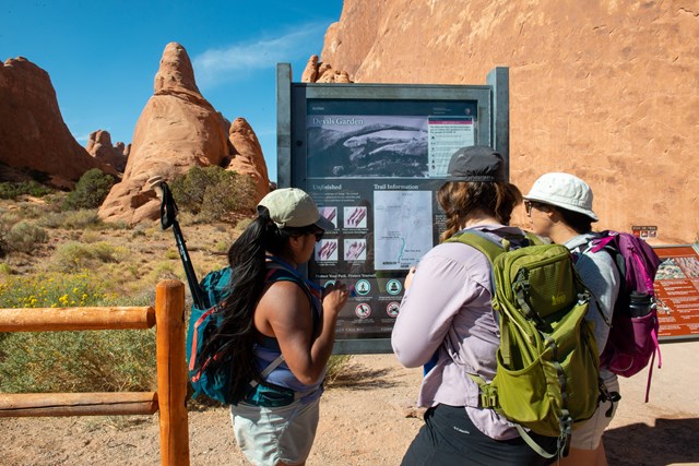Hikers reading a trailhead sign near a fence, sandstone walls in the background