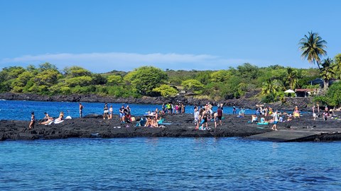 Snorkelers gather on the black lava rock shoreline among the blue waters of Hōnaunau Bay.