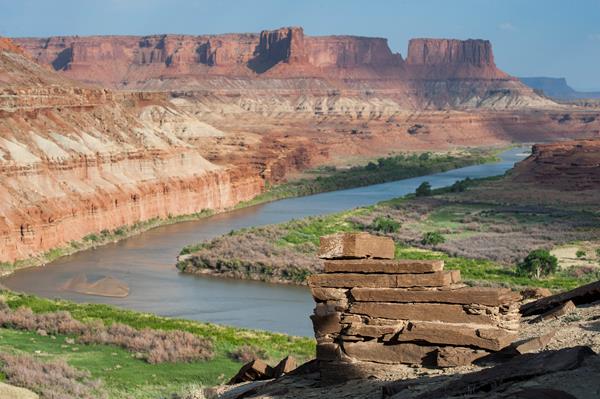 Stacked sandstone slabs sit atop a cliff overlooking the green river.