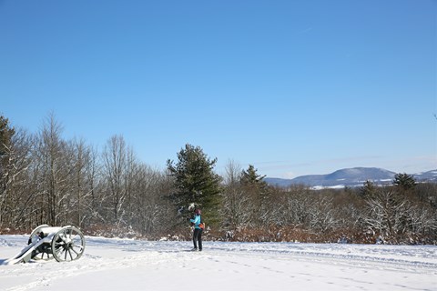 A cross-country skier traverses down a groomed path on a hilltop and past a cannon covered in snow.