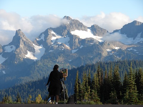 A female figure viewed from behind stands at an easel painting a picture of the tall mountains in fr
