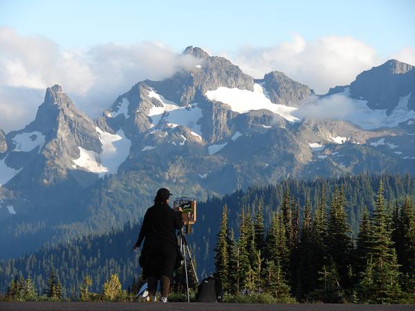 A female figure viewed from behind stands at an easel painting a picture of the tall mountains in fr