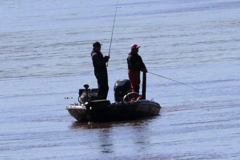 Two men with fishing poles standing on a small boat in the water,