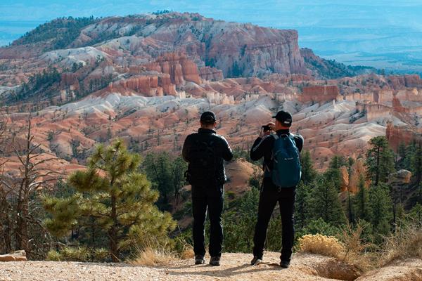 Two hikers pause along a path to view a red rock landscape of badlands and spires at a cliff's edge