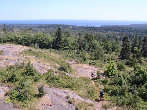 Two people with backpacks walk down a trail on a ridge surrounded by forest.