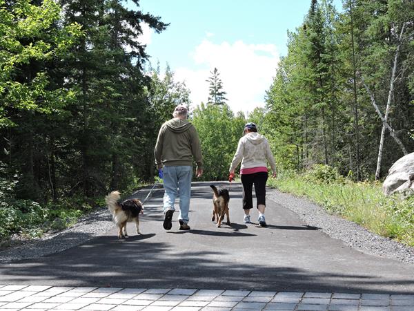 Two visitors walk along the Rainy Lake Recreation Trail, each one with a dog on a leash.