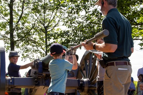 A child stands near a cannon near a park volunteer who instructs him on its use.