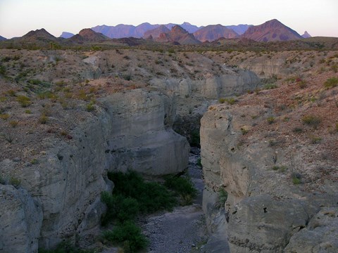 The setting sun casts a glow over a canyon composed of gray tuff, with mountains rising behind.