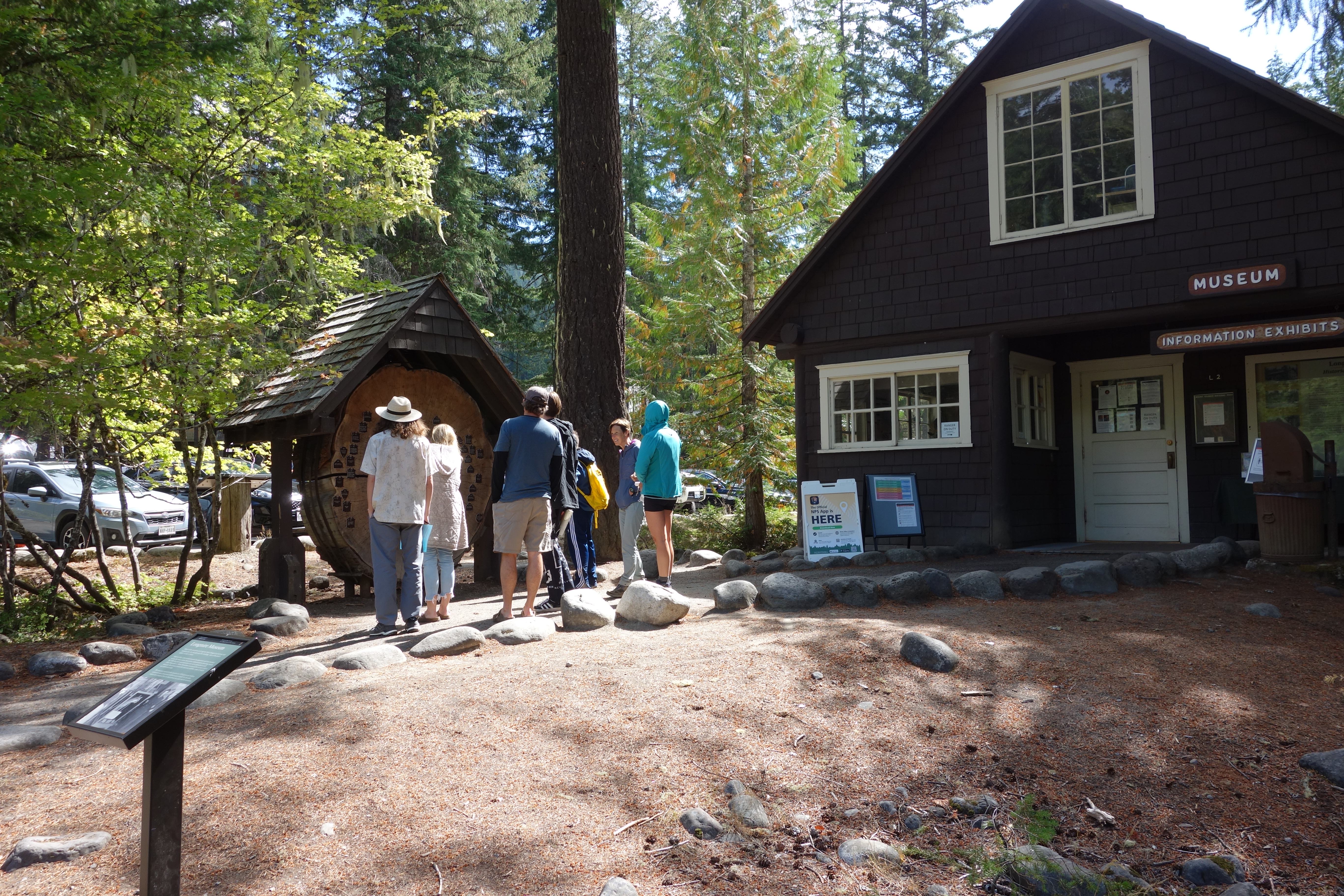 A small wayside panel in front of a wood cabin. Visitors stand on a path next to the cabin.