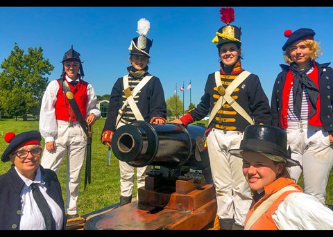 6 people dressed as soldier, marine, and sailors from the War of 1812 pose for a picture around a re