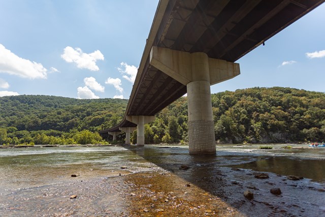 The US-340 highway bridge crossing the river high overhead, seen from the rocky river shoreline below
