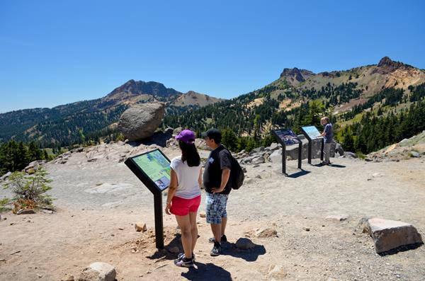 Visitors standing reading waysides about geology in the Bumpass Hell parking area with the glacial e