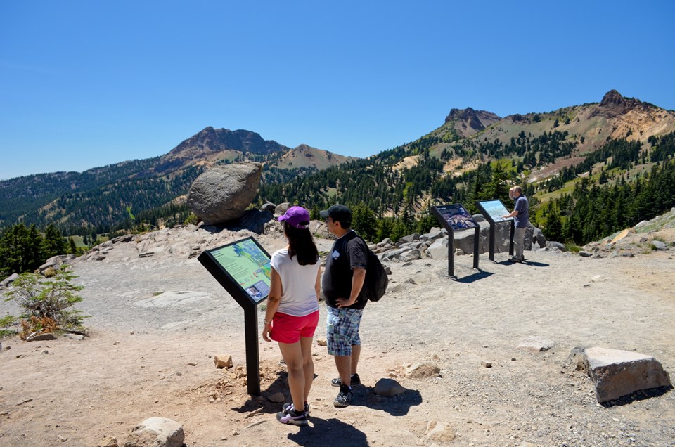 Visitors standing reading waysides about geology in the Bumpass Hell parking area with the glacial e