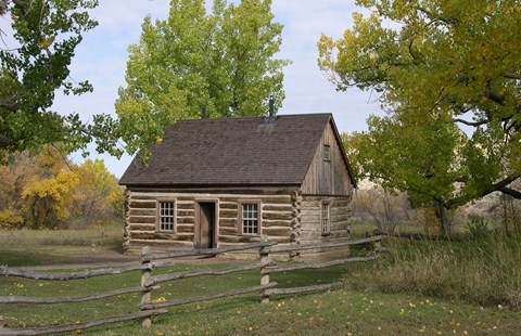A log cabin sits in a grassy area on the other side of a wooden fence, trees and blue sky behind it.