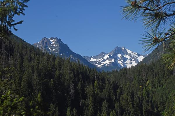 Two gray mountain peaks rise from a forested valley. One mountain is partially covered in glaciers