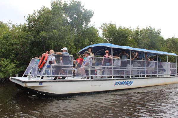 A large open air white boat with blue canopy top with passengers seated motors through murky water
