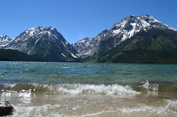 Waves crash on the shore of a clear, blue lake at the base of a mountain range.