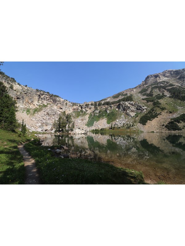 An alpine lake sits at the base of a rocky cliff surrounded by green vegetation.