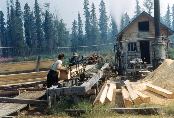 Two men working on sawmill stacked lumber in foreground and background.
