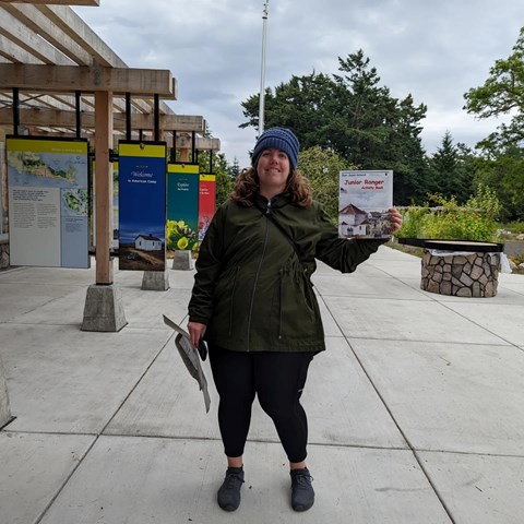femme looking person in a coat and hat holds up a junior ranger booklet and is smiling