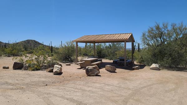 Three picnic tables and a shade structure surrounded by desert vegetation.
