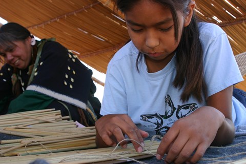 A color photo of a young girl leaning forward over a table and weaving rope around dried tule reeds.
