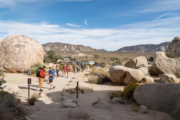 A group of hikers heading up a dirt trail between large rocks with mountains in the background.