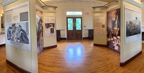Exhibit panels with text and images are mounted on free-standing walls. wood-paneled floor