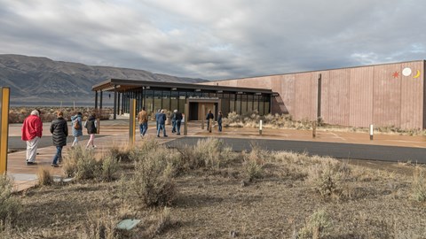 Color photograph of a building made of red stone blocks with a glassy entranceway with many people e