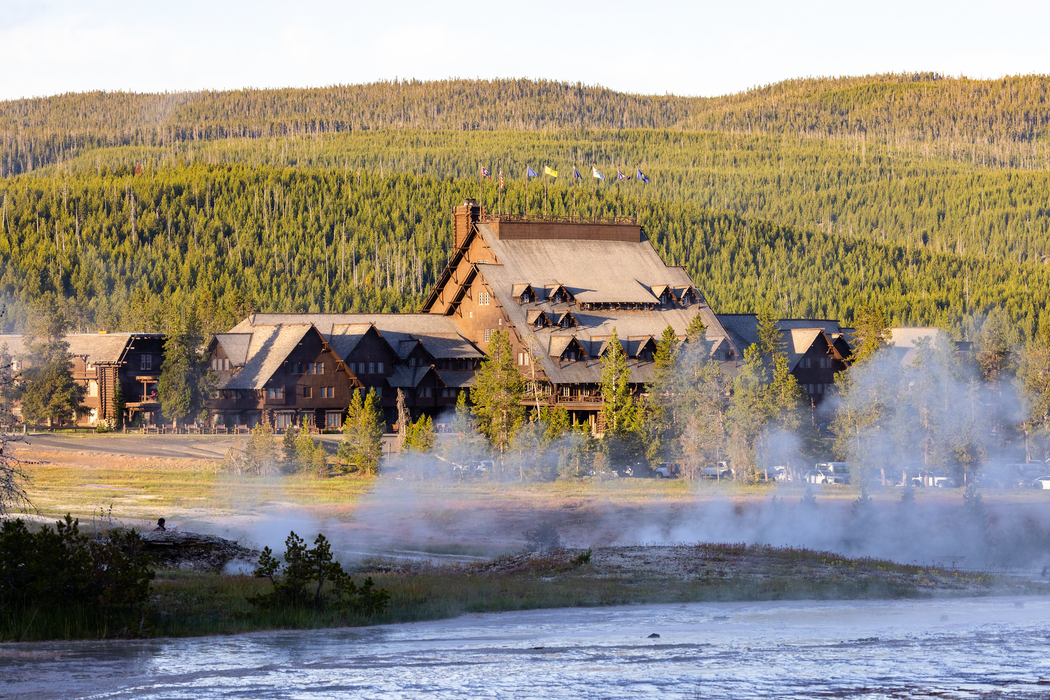 The large Old Faithful Inn with a steep, shingled roofline and flags on top, seen across a steaming thermal area with forested hills behind.