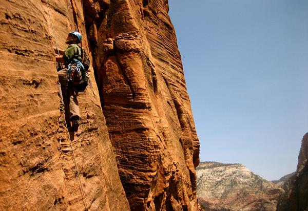 Red-orange sandstone cliff-face with a rock climber in a helmet making his way up.