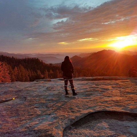 A woman stands on a granite dome watching the sunset.